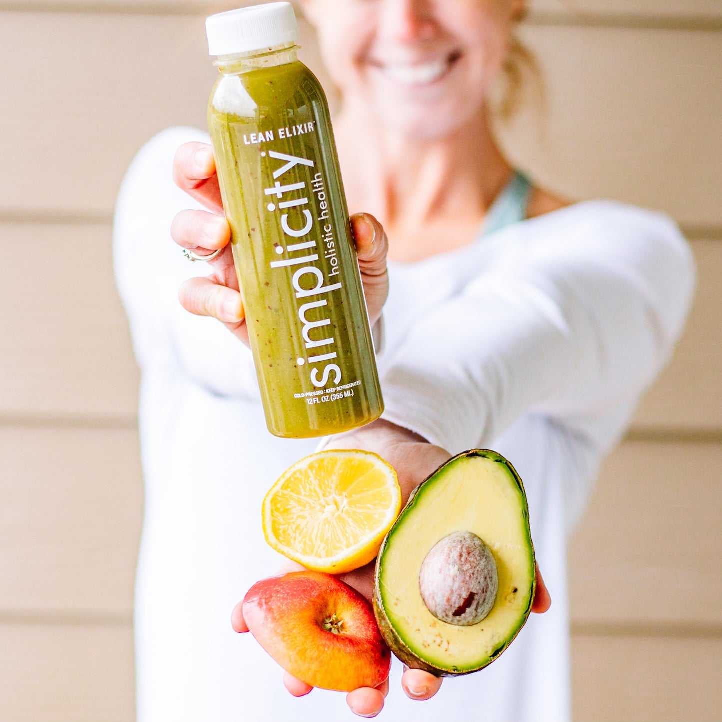 Athletic woman holding bottle of Simplicity Lean Elixir, an apple, a lemon, and an avocado.